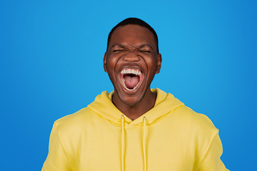An exultant african american man in a yellow hoodie laughs heartily with his mouth wide open, eyes closed in joy, against a crisp blue background, embodying pure elation