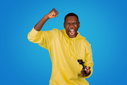 An exultant Black man in a yellow hoodie clenches his fist in a gesture of victory and shouts in excitement while holding a game controller, against a bright blue background