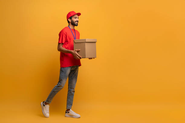 istock Smiling delivery man in red uniform and cap carrying a cardboard box 2000901407