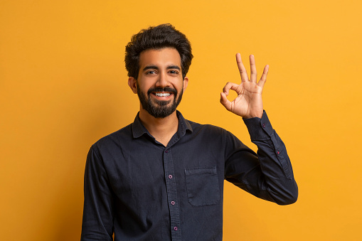 Young smiling indian man making 'OK' gesture at camera, joyful handsome eastern guy gesturing okay, cheerful male showing sign of approval while standing against yellow background, copy space