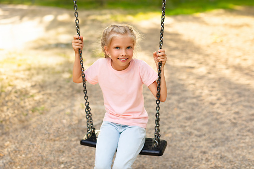 Happy schoolgirl sitting on swing while walking on playground, girl looking and smiling at camera, copy space. Outdoor summer activities for children