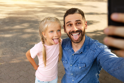 Funny middle aged father walking with his little daughter outdoors in park and making selfie by mobile phone, grimacing and showing tongues to phone camera