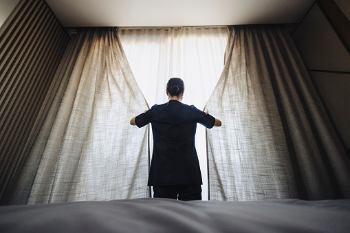 Rare view of uniformed hotel maid opening the white curtains in hotel room