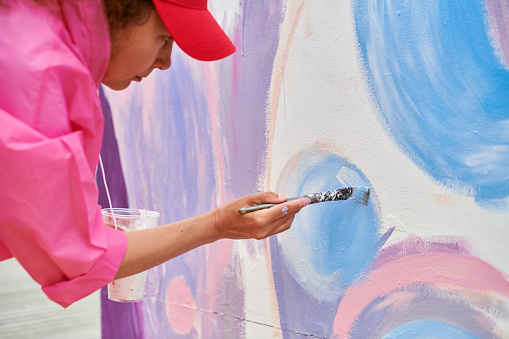 Female painter in cap draws picture with paintbrush on canvas for outdoor street exhibition, close up side view of female artist apply brushstrokes to canvas, symphony of art creativity