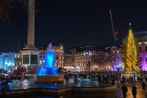 A magical evening in Trafalgar Square, London, with a bluelit fountain, bustling crowd, and a grand Christmas tree adorned with golden lights, set against iconic landmarks.