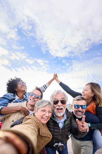 Vertical group diverse funny mature tourist friends posing piggyback with hands joined in air taking selfie together with front camera on travel outdoor. Adult happy six excited people enjoy holidays