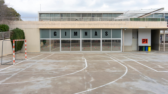 An empty outdoor sports court awaits players, with a gym's large glass facade reflecting the open sky in majorca