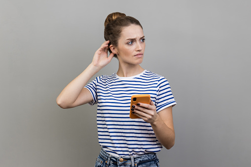 Portrait of pensive thoughtful woman wearing striped T-shirt standing with mobile phone, touching head, having doubt suspicion feeling. Indoor studio shot isolated on gray background.