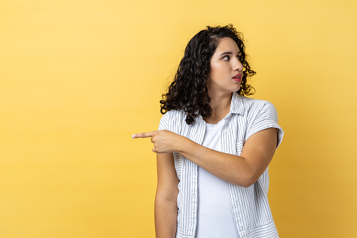 Portrait of woman with dark wavy hair pointing finger aside, ordering get out and looking resentful, boss dismissing from work, showing exit. Indoor studio shot isolated on yellow background.
