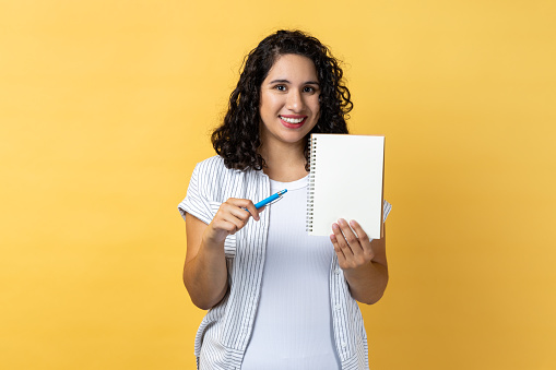 Portrait of joyful smiling positive woman with dark wavy hair standing holding paper notebook and showing empty paper, copy space. Indoor studio shot isolated on yellow background.