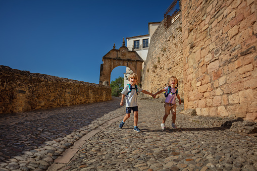 Happy kids with backpacks run in paved streets of Ronda, enjoy their summer vacations in the South of Spain, Europe