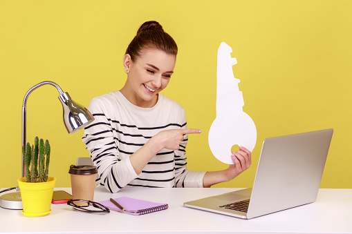 Woman office worker pointing at big paper key and looking at laptop screen with toothy smile while sitting on workplace, house purchase concept. Indoor studio studio shot isolated on yellow background