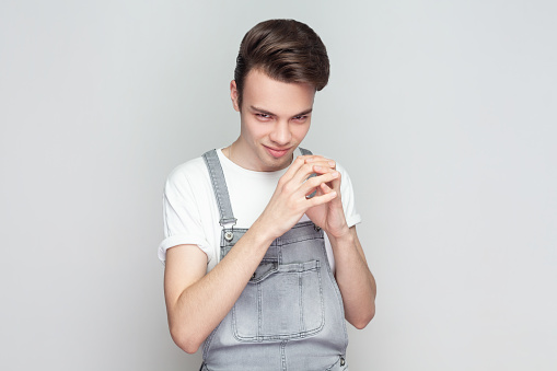 My plan is perfect. Young brunette man schemes something, steepls fingers and looks with cunning expression aside, smiles sly, wearing denim overalls. Indoor studio shot isolated on gray background.