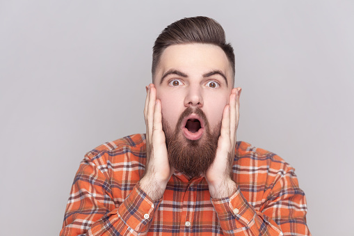 Portrait of shocked amazed bearded man looking at camera with big eyes and open mouth, keeps palms on cheeks, wearing checkered shirt. Indoor studio shot isolated on gray background.