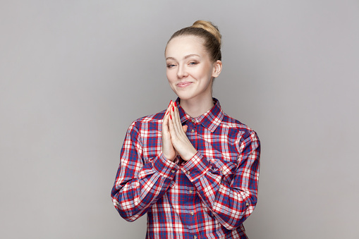 Portrait of sly attractive woman with bun hairstyle keeps palms together, planning devil plan or prank, wearing checkered shirt. Indoor studio shot isolated on gray background.