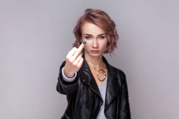 Serious rude woman standing showing middle finger, impolite gesture, looking at camera. Portrait of serious rude woman with short hairstyle standing showing middle finger, impolite gesture, looking at camera, wearing black leather jacket. Indoor studio shot isolated on grey background. asshole stock pictures, royalty-free photos & images