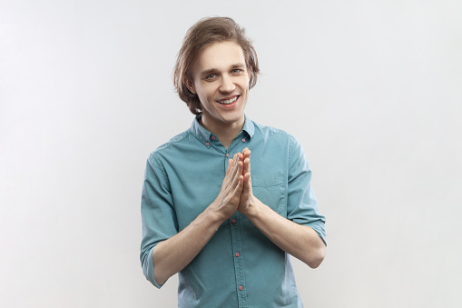 Portrait of cunning young handsome man rubbing palm as having cunning evil idea, devious plan in mind, thinking revenge, wearing blue shirt. Indoor studio shot isolated on gray background.