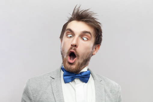 Portrait of funny crazy childish bearded man keeps mouth open and eyes crossed, making foolish face, wearing grey suit and blue bow tie. Indoor studio shot isolated on gray background.
