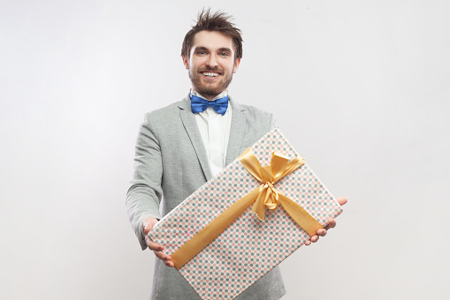 Portrait of positive optimistic smiling bearded man standing holding present box, giving gift to wife on anniversary, wearing grey suit and blue bow tie. Indoor studio shot isolated on gray background