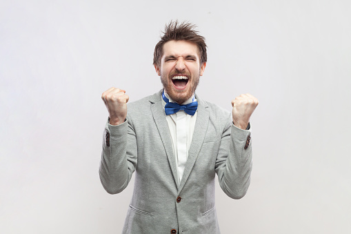 Portrait of extremely happy overjoyed bearded man screaming happiness, clenched fists, celebrating his victory, wearing grey suit and blue bow tie. Indoor studio shot isolated on gray background.