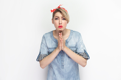 Portrait of sad hopeful blonde woman wearing blue denim shirt and red headband standing with praying gesture, asking to forgive her. Indoor studio shot isolated on gray background.