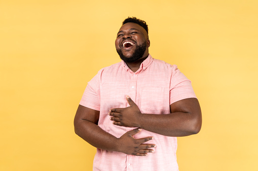 Portrait of happy positive funny bearded man wearing pink shirt laughing out loud, holding his belly, hearing funny joke, being in good mood. Indoor studio shot isolated on yellow background.