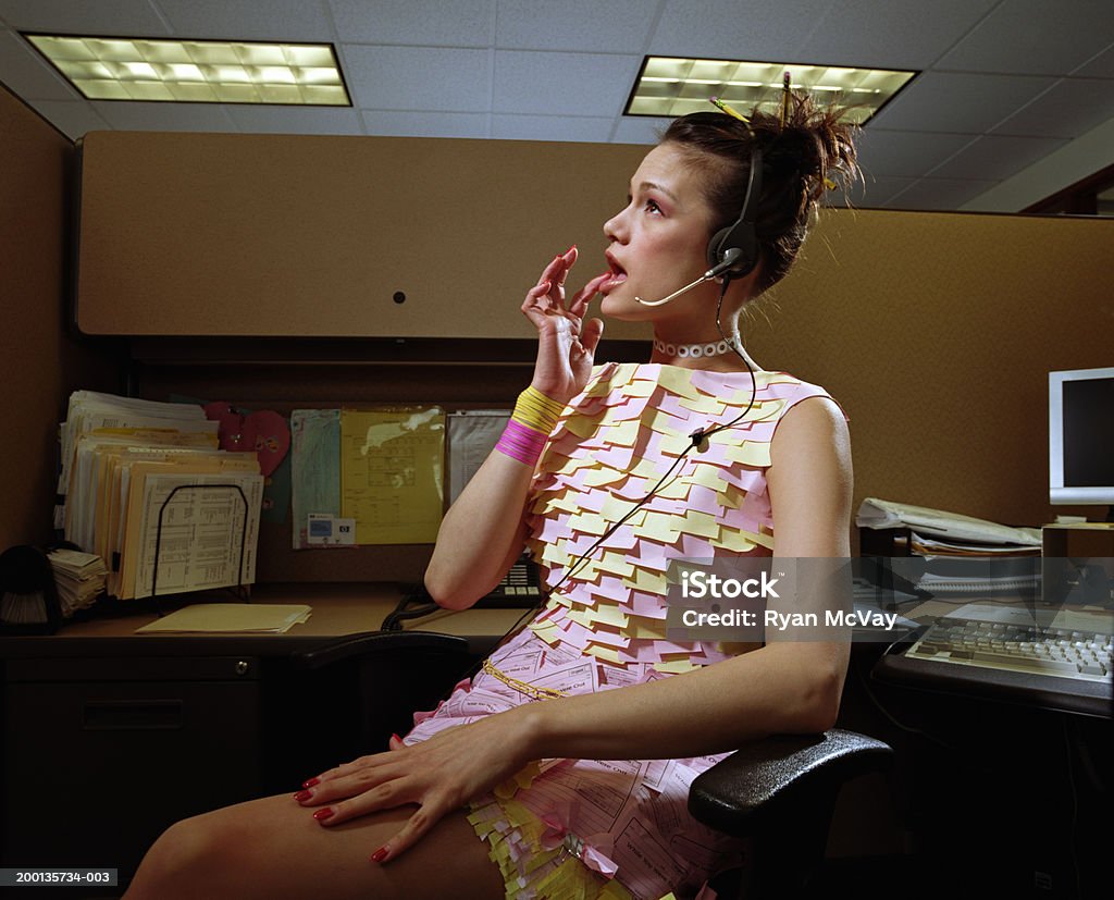 Woman in dress made of office supplies, at desk, wearing headset  Wasting Time Stock Photo
