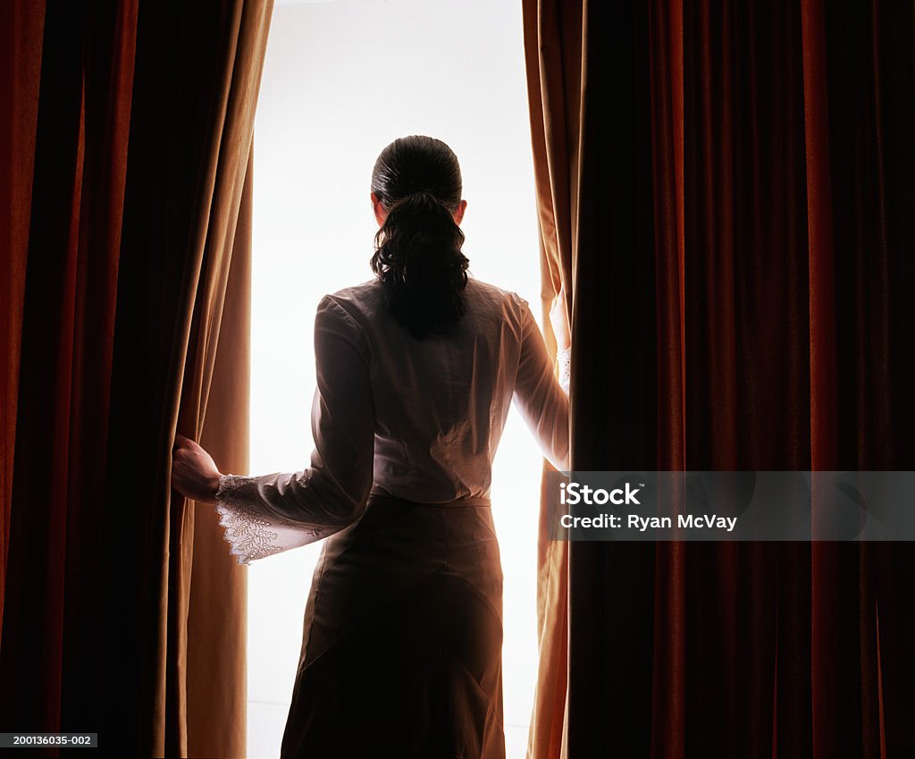Young woman looking through red curtains, rear view  Mystery Stock Photo