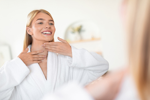Young blonde woman in bathrobe gently applies moisturizer to her neck and face skin in bathroom interior, smiling to her reflection in mirror, enjoying healthy skincare and pampering