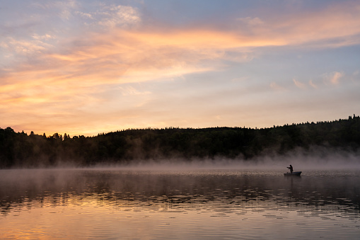 A senior man doing fly-fishing in a boat on a lake of Lanaudiere, Quebec, during a beautiful sunrise of summer.