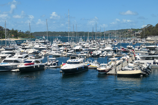 Nautical vessels and real estate worth hundreds of millions of dollars at Middle Harbour, Sydney.  In the foreground is Middle Harbour Marina.  In the distance are residential districts: on the left is Balgowlah; and in the centre and on the right is Mosman. This image was taken from the Spit Bridge on a hot and sunny afternoon on 10 February 2024.