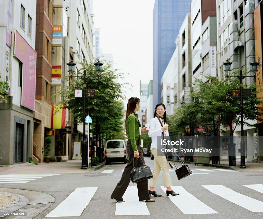 Dos mujeres jóvenes crossing urban street, tener una conversación, lateral - Foto de stock de Andar libre de derechos
