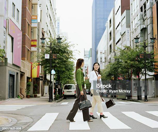 Dos Mujeres Jóvenes Crossing Urban Street Tener Una Conversación Lateral Foto de stock y más banco de imágenes de Andar