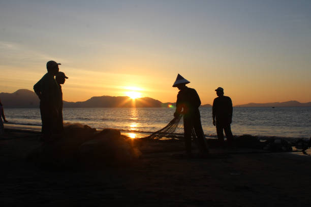 Silhouette fishermen are cleaning their nets and collecting fish on the beach at sunset. In the reddish-orange light of dusk, they diligently cleaned the nets of the day's catch. A peaceful atmosphere is created amidst the sparkling sea, while the sky welcomes the night with charming warmth  07/23/2017