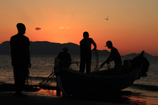 Silhouette fishermen are cleaning their nets and collecting fish on the beach at sunset. In the reddish-orange light of dusk, they diligently cleaned the nets of the day's catch. A peaceful atmosphere is created amidst the sparkling sea, while the sky welcomes the night with charming warmth  07/23/2017