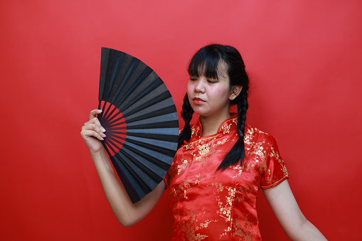 An Asian teenage girl in a Chinese traditional red dress (Cheongsam or Qipao), holding a black hand fan posted in the studio Isolated on a red background.