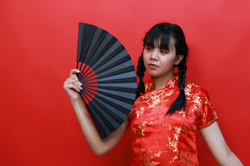 An Asian teenage girl in a Chinese traditional red dress (Cheongsam or Qipao), holding a black hand fan posted in the studio Isolated on a red background.