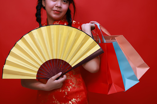 Happy Cute Young Asian shopper girl wearing a traditional cheongsam qipao dress holding a yellow hand fan and shopping bag isolated on a red background. Happy Chinese New Year