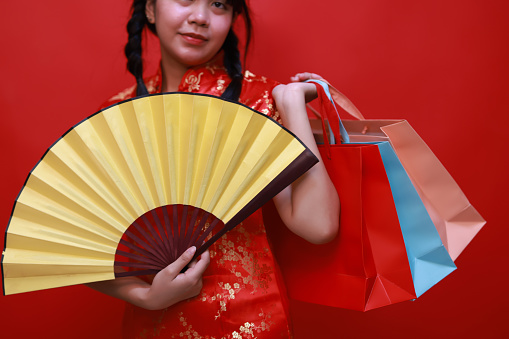 Happy Cute Young Asian shopper girl wearing a traditional cheongsam qipao dress holding a yellow hand fan and shopping bag isolated on a red background. Happy Chinese New Year