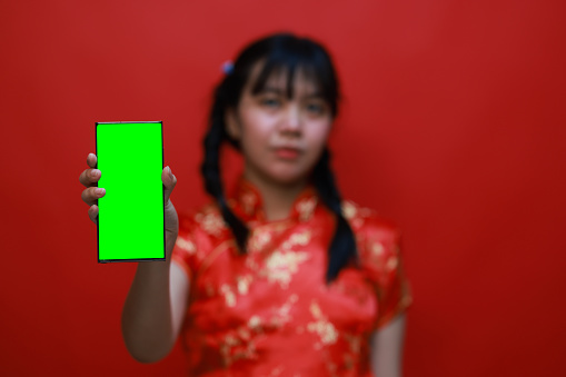 A cute Young Asia girl wearing a traditional cheongsam dress called qipao holds a mobile phone with a green screen on Chinese New Year's Day