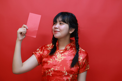 A lovely girl looking & smiling joyfully at a red envelope in Chinese New Year, an Asian woman wearing a Chinese dress, holding a red envelope, glad to receive money as a New Year gift.