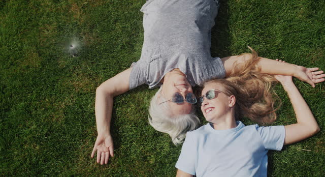 Grandmother and granddaughter lie on the grass under jets of water, laugh and escape from the heat. Slow motion video