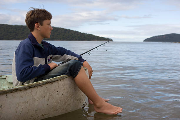 Boy (12-14) fishing from boat, feet hanging over side stock photo