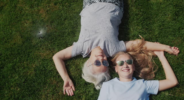 Senior woman and granddaughter lie on the grass under jets of water, laugh and escape from the heat. Slow motion video