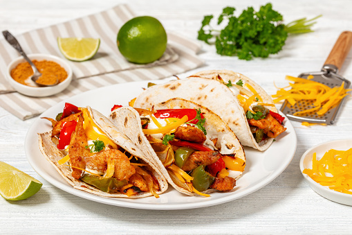 close-up of baked tex-mex chicken fajitas with mixed sweet pepper, onion, sour cream, shredded cheese, and white corn tortillas on white plate on wooden table with ingredients, landscape view