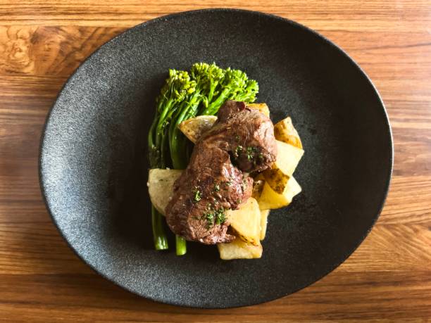 Horizontal high angle closeup photo of a pan fried Australian Eye Fillet steak served with steamed green broccoli and roast potatoes on a black ceramic plate on a wooden table in a fine dining restaurant. Uralla, New England high country NSW.
