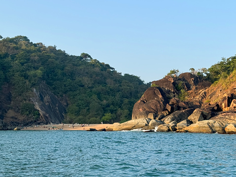 Stock photo showing the sea and coastline rocks off Honeymoon Island, Goa on a sunny day with clear blue skies.  Vacationing holidaymakers sunbathing on golden sandy beach reached by boat.