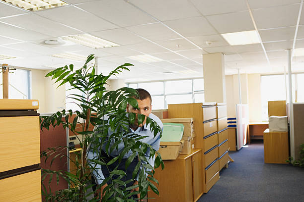 man hiding behind plant in office looking through foliage - hiding fear men peeking photos et images de collection