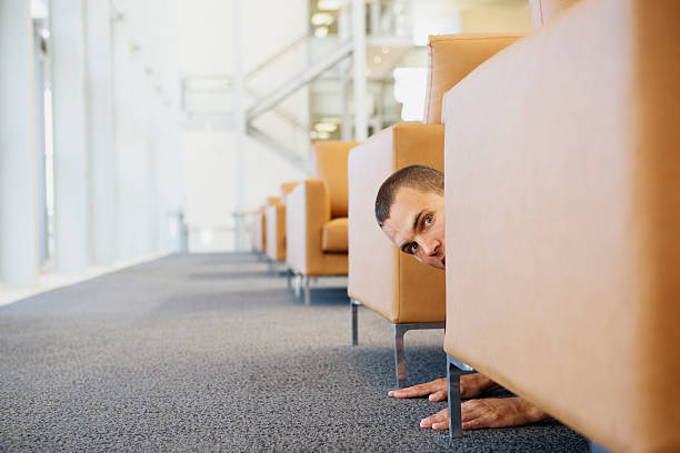 man crawling on floor peering around sofa - hiding fear men peeking fotografías e imágenes de stock