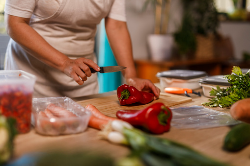 Cut out shot of diligent homemaker, wearing an apron, standing over a kitchen table, chopping and preparing various colorful vegetables for winter preservation.